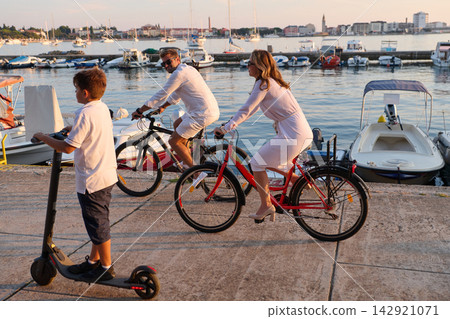 Family Cycling Along Waterfront Marina At Sunset With Child On Scooter And Boats Nearby Family Cycling Along Waterfront Marina At Sunset With Child On Scooter And Boats Nearby 142921071