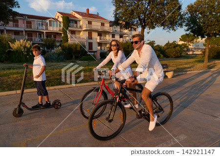 Family Biking And Scooter Ride On Seaside Promenade During Golden Hour Summer Vacation Family Biking And Scooter Ride On Seaside Promenade During Golden Hour Summer Vacation 142921147