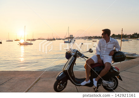 Man on Scooter at Sunset by Harbor With Boats Relaxing on a Seaside Summer Vacation Man on Scooter at Sunset by Harbor With Boats Relaxing on a Seaside Summer Vacation 142921161