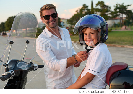Father Helping Son Fasten Helmet On Scooter During Summer Evening Ride Near Park Father Helping Son Fasten Helmet On Scooter During Summer Evening Ride Near Park 142921354