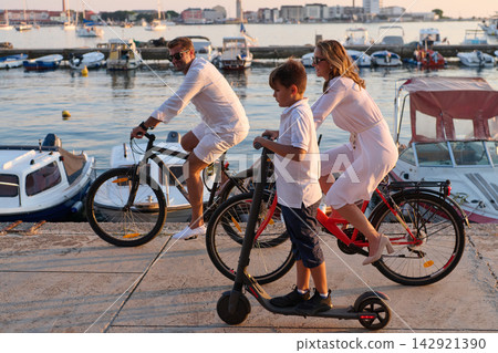 Family Riding Bicycles and Electric Scooter Along Waterfront Harbor at Sunset in Summer Vacation Family Riding Bicycles and Electric Scooter Along Waterfront Harbor at Sunset in Summer Vacation 142921390