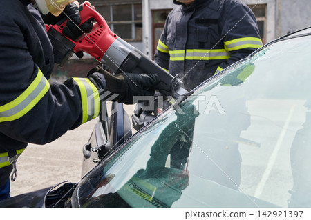 Firefighters Performing Vehicle Extrication Using Power Saw To Remove Cracked Windshield After Car Accident Firefighters Performing Vehicle Extrication Using Power Saw To Remove Cracked Windshield After Car Accident 142921397