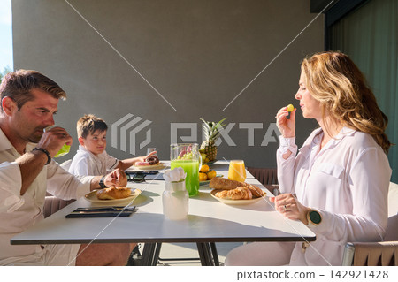 Family Enjoying Outdoor Breakfast on Terrace With Croissants, Fresh Juice and Relaxed Weekend Vibe Family Enjoying Outdoor Breakfast on Terrace With Croissants, Fresh Juice and Relaxed Weekend Vibe 142921428