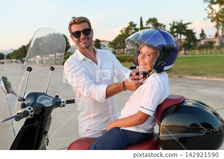 Father Helps Son Fasten Helmet on Scooter During Sunny Coastal Family Outing Father Helps Son Fasten Helmet on Scooter During Sunny Coastal Family Outing 142921590