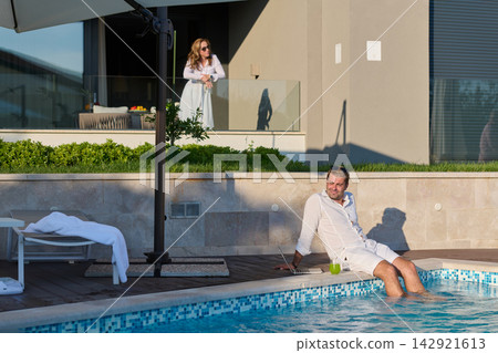 Man Relaxing by Pool With Laptop While Woman Watches From Balcony at Luxury Vacation Home Man Relaxing by Pool With Laptop While Woman Watches From Balcony at Luxury Vacation Home 142921613