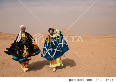 Two Egyptian whirling dancers wearing vivid embroidered traditional costumes strike theatrical poses on the Giza desert plateau with the iconic Two Egyptian whirling dancers wearing vivid embroidered traditional costumes strike theatrical poses on the Giza desert plateau with the iconic 142921643