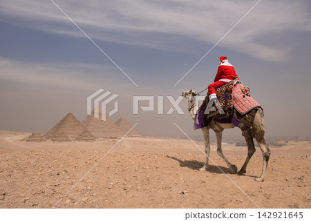 Santa claus riding a decorated camel across the arid Giza desert plateau with the ancient pyramids in the misty background Santa claus riding a decorated camel across the arid Giza desert plateau with the ancient pyramids in the misty background 142921645