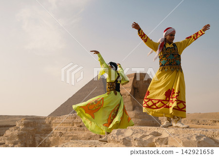 Two exuberant folk dancers wearing vivid embroidered yellow robes leap and swirl with arms raised beside the Great Sphinx and Great Pyramid of Giza Two exuberant folk dancers wearing vivid embroidered yellow robes leap and swirl with arms raised beside the Great Sphinx and Great Pyramid of Giza 142921658