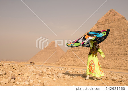 Egyptian folk dancer in neon yellow embroidered robe twirling a dramatic billowing multicolored cape before the iconic Giza Pyramids on sandy desert Egyptian folk dancer in neon yellow embroidered robe twirling a dramatic billowing multicolored cape before the iconic Giza Pyramids on sandy desert 142921660