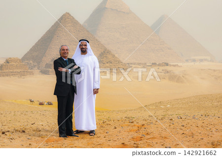 Multicultural business partnership portrait of Arab man in thobe and senior Western executive smiling together at Giza Pyramids Egypt desert ideal for Multicultural business partnership portrait of Arab man in thobe and senior Western executive smiling together at Giza Pyramids Egypt desert ideal for 142921662
