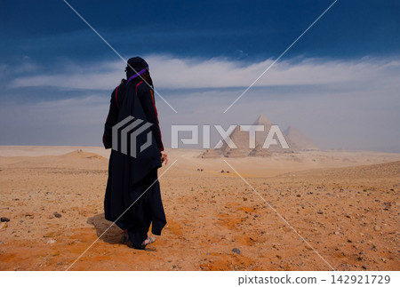 Solitary woman in black abaya gazing at iconic Giza Pyramids across vast Egyptian desert plateau under dramatic blue sky ideal for travel cultural Solitary woman in black abaya gazing at iconic Giza Pyramids across vast Egyptian desert plateau under dramatic blue sky ideal for travel cultural 142921729