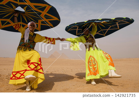 Two Egyptian tanoura folk dancers in vivid embroidered robes hold hands spinning colorful skirts overhead at the Giza Pyramids in Egypt ideal for Two Egyptian tanoura folk dancers in vivid embroidered robes hold hands spinning colorful skirts overhead at the Giza Pyramids in Egypt ideal for 142921735