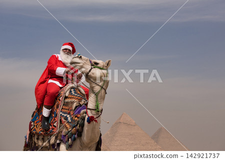 Cheerful Santa Claus in full red costume leans forward affectionately stroking decorated riding camel with ancient pyramid backdrop in hazy Egyptian Cheerful Santa Claus in full red costume leans forward affectionately stroking decorated riding camel with ancient pyramid backdrop in hazy Egyptian 142921737