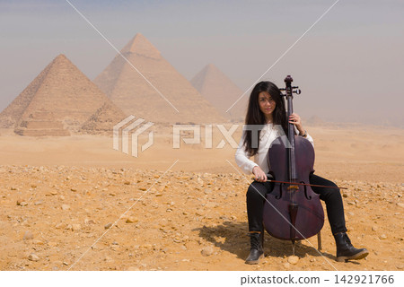 Young female cellist seated playing cello with iconic Giza Pyramids backdrop in Egyptian desert ideal for arts travel and cultural campaigns Young female cellist seated playing cello with iconic Giza Pyramids backdrop in Egyptian desert ideal for arts travel and cultural campaigns 142921766
