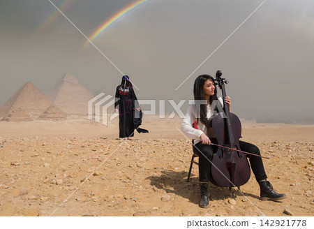 Female cellist seated performing cello at iconic Giza Pyramids with traditionally dressed woman and vivid rainbow arc in dramatic desert sky ideal for Female cellist seated performing cello at iconic Giza Pyramids with traditionally dressed woman and vivid rainbow arc in dramatic desert sky ideal for 142921778