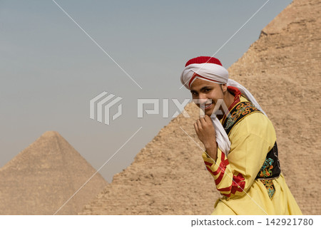 Authentic portrait of a joyful young cultural performer in ornate yellow Arab folk dress adjusting turban headwrap while smiling at camera against Authentic portrait of a joyful young cultural performer in ornate yellow Arab folk dress adjusting turban headwrap while smiling at camera against 142921780