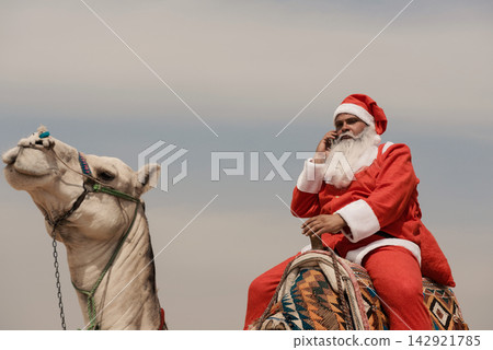 Santa Claus in full red costume riding white camel while talking on cell phone under overcast desert sky a bold multicultural holiday humor concept 142921785