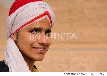 Authentic egyptian portrait of a charming young man in a traditional red and white turban smiling with direct eye contact outdoors against a warm Authentic egyptian portrait of a charming young man in a traditional red and white turban smiling with direct eye contact outdoors against a warm 142921787