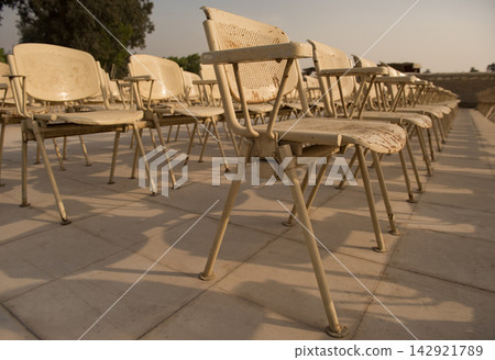 Rows of desolate empty chairs on a sun warmed tiled outdoor plaza with peeling paint and rust details receding into soft bokeh ideal for absence Rows of desolate empty chairs on a sun warmed tiled outdoor plaza with peeling paint and rust details receding into soft bokeh ideal for absence 142921789