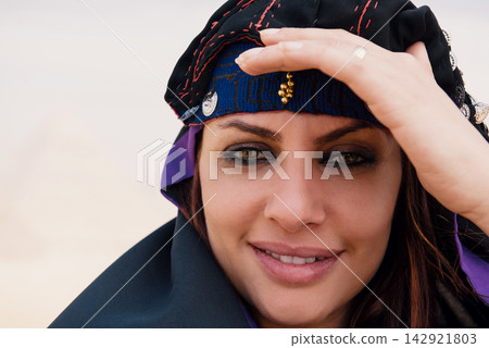 Arab woman wearing an ornate embroidered coin headdress and dark abaya smiles warmly in an intimate close up portrait with a blurred sandy desert Arab woman wearing an ornate embroidered coin headdress and dark abaya smiles warmly in an intimate close up portrait with a blurred sandy desert 142921803