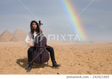 Young Arab female cellist performing under a dramatic natural rainbow at iconic Giza pyramids in Egyptian desert ideal for music arts and cultural Young Arab female cellist performing under a dramatic natural rainbow at iconic Giza pyramids in Egyptian desert ideal for music arts and cultural 142921805