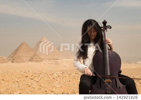 Female cellist deeply absorbed in playing cello outdoors on the Giza plateau with three ancient Egyptian pyramids in the hazy desert background ideal Female cellist deeply absorbed in playing cello outdoors on the Giza plateau with three ancient Egyptian pyramids in the hazy desert background ideal 142921826