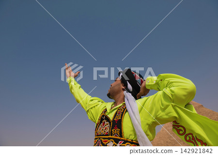 Triumphant Egyptian folk dancer in vibrant neon yellow embroidered robe and turban raises arms skyward in dramatic low angle shot with ancient Giza Triumphant Egyptian folk dancer in vibrant neon yellow embroidered robe and turban raises arms skyward in dramatic low angle shot with ancient Giza 142921842