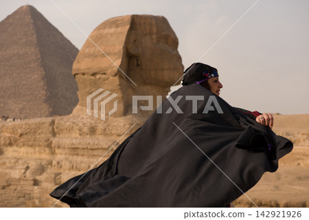 Sphinx portrait of a proud Arab woman in embroidered black abaya and ornamental headpiece standing before Great Sphinx of Giza with pyramid softly Sphinx portrait of a proud Arab woman in embroidered black abaya and ornamental headpiece standing before Great Sphinx of Giza with pyramid softly 142921926