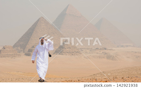 Smiling arab traveler in traditional white kandura strides across sandy Giza plateau with three ancient pyramids in hazy background ideal for MENA Smiling arab traveler in traditional white kandura strides across sandy Giza plateau with three ancient pyramids in hazy background ideal for MENA 142921938