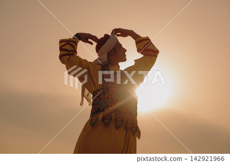 Traditional folk dancer in embroidered Egyptian heritage costume performing arms raised pose against warm golden sunset sky with dramatic lens flare Traditional folk dancer in embroidered Egyptian heritage costume performing arms raised pose against warm golden sunset sky with dramatic lens flare 142921966