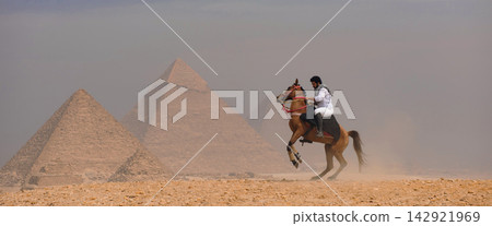 Arab horseman controlling rearing stallion on sandy giza plateau with three iconic pyramids rising through dusty haze dramatic egypt travel and Arab horseman controlling rearing stallion on sandy giza plateau with three iconic pyramids rising through dusty haze dramatic egypt travel and 142921969