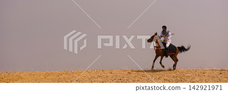 Proud arab horseman in traditional white thobe and keffiyeh riding arabian horse at full gallop across an arid rocky desert ridge with dramatic hazy Proud arab horseman in traditional white thobe and keffiyeh riding arabian horse at full gallop across an arid rocky desert ridge with dramatic hazy 142921971
