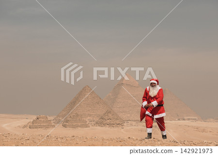 Iconic pyramid backdrop frames a surreal and festive Santa Claus in full red costume holding a gift bag on the sandy Giza plateau under a hazy Iconic pyramid backdrop frames a surreal and festive Santa Claus in full red costume holding a gift bag on the sandy Giza plateau under a hazy 142921973