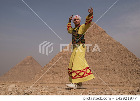 Expressive young Middle Eastern folk dancer in vibrant yellow embroidered robe raising arms joyfully before the iconic Great Pyramids of Giza on the Expressive young Middle Eastern folk dancer in vibrant yellow embroidered robe raising arms joyfully before the iconic Great Pyramids of Giza on the 142921977