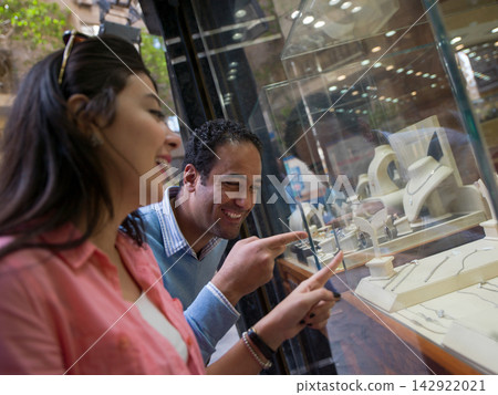 Happy couple shopping for jewelry at a store window, smiling and pointing at necklace display Happy couple shopping for jewelry at a store window, smiling and pointing at necklace display 142922021