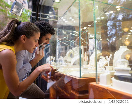 Couple window shopping for jewelry at a boutique display, smiling and pointing at rings and necklaces 142922031