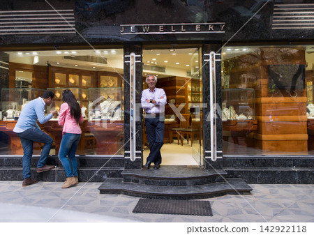 Jewellery shop owner standing at the entrance while customers inspect display window on city street Jewellery shop owner standing at the entrance while customers inspect display window on city street 142922118