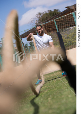Male athlete working out with battle ropes outdoors at an open-air CrossFit style fitness gym with a friend Male athlete working out with battle ropes outdoors at an open-air CrossFit style fitness gym with a friend 142922282
