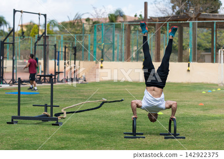 Male athlete performing handstand push-up on parallettes at outdoor CrossFit gym with friend Male athlete performing handstand push-up on parallettes at outdoor CrossFit gym with friend 142922375