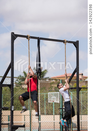 Two men climbing ropes at an outdoor CrossFit gym during partner fitness training session Two men climbing ropes at an outdoor CrossFit gym during partner fitness training session 142922398