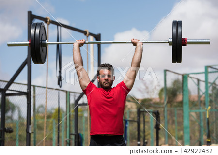 Middle Eastern man lifting barbell overhead in outdoor CrossFit gym, focused strength training session Middle Eastern man lifting barbell overhead in outdoor CrossFit gym, focused strength training session 142922432