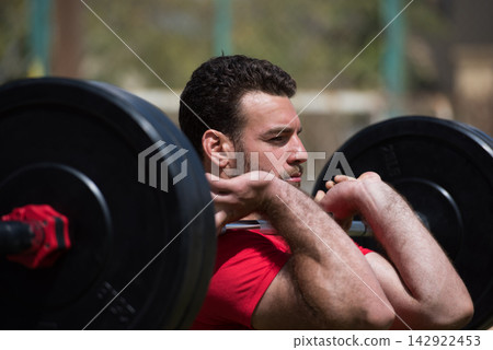 Arab man performing front squat with barbell in an outdoor CrossFit gym with a friend Arab man performing front squat with barbell in an outdoor CrossFit gym with a friend 142922453