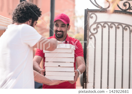 Friendly delivery driver handing stacked pizza boxes to customer at home gate during festive holiday dinner Friendly delivery driver handing stacked pizza boxes to customer at home gate during festive holiday dinner 142922681