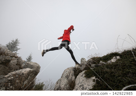 Woman In Red Jacket Leaping Between Rocky Ledges During Foggy Mountain Hiking Adventure 142922985