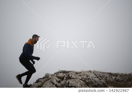 Trail Runner Wearing Headlamp Climbing Rocky Hill in Foggy Winter Landscape at Dawn Trail Runner Wearing Headlamp Climbing Rocky Hill in Foggy Winter Landscape at Dawn 142922987