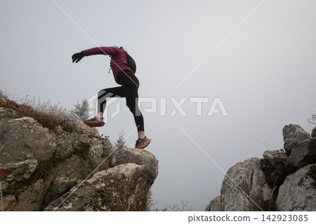 Woman Hiker Jumping Across Rocky Gap On Foggy Mountain Trail With Backpack And Sport Shoes Woman Hiker Jumping Across Rocky Gap On Foggy Mountain Trail With Backpack And Sport Shoes 142923085