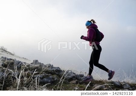 Woman Trail Running on Frosty Mountain Ridge at Sunrise Wearing Winter Jacket and Headband Woman Trail Running on Frosty Mountain Ridge at Sunrise Wearing Winter Jacket and Headband 142923110