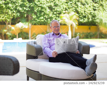 Middle Eastern senior man reading newspaper and relaxing at a modern luxury villa by the pool Middle Eastern senior man reading newspaper and relaxing at a modern luxury villa by the pool 142923239