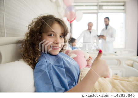 Smiling girl gives thumbs up in Middle Eastern eye hospital ward with doctors in background 142923846