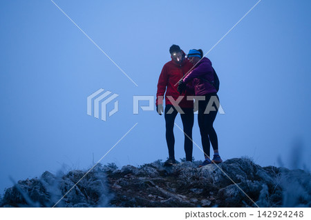 Couple Hiking On Mountain Summit At Dawn Wearing Headlamp And Winter Jackets Couple Hiking On Mountain Summit At Dawn Wearing Headlamp And Winter Jackets 142924248